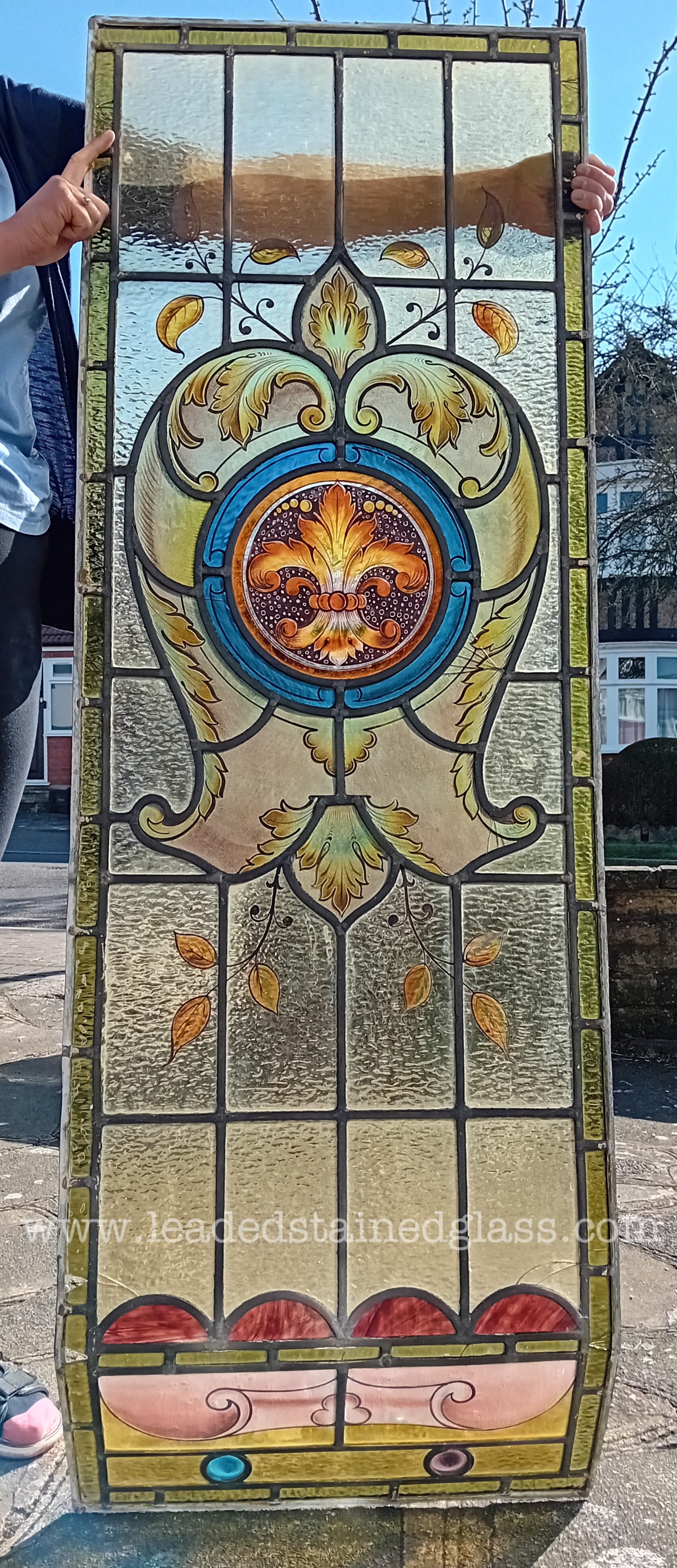 A hand painted leaded stained glass window panel with a kink at the bottom. It features a fleur-de-lis roundel in the middle.