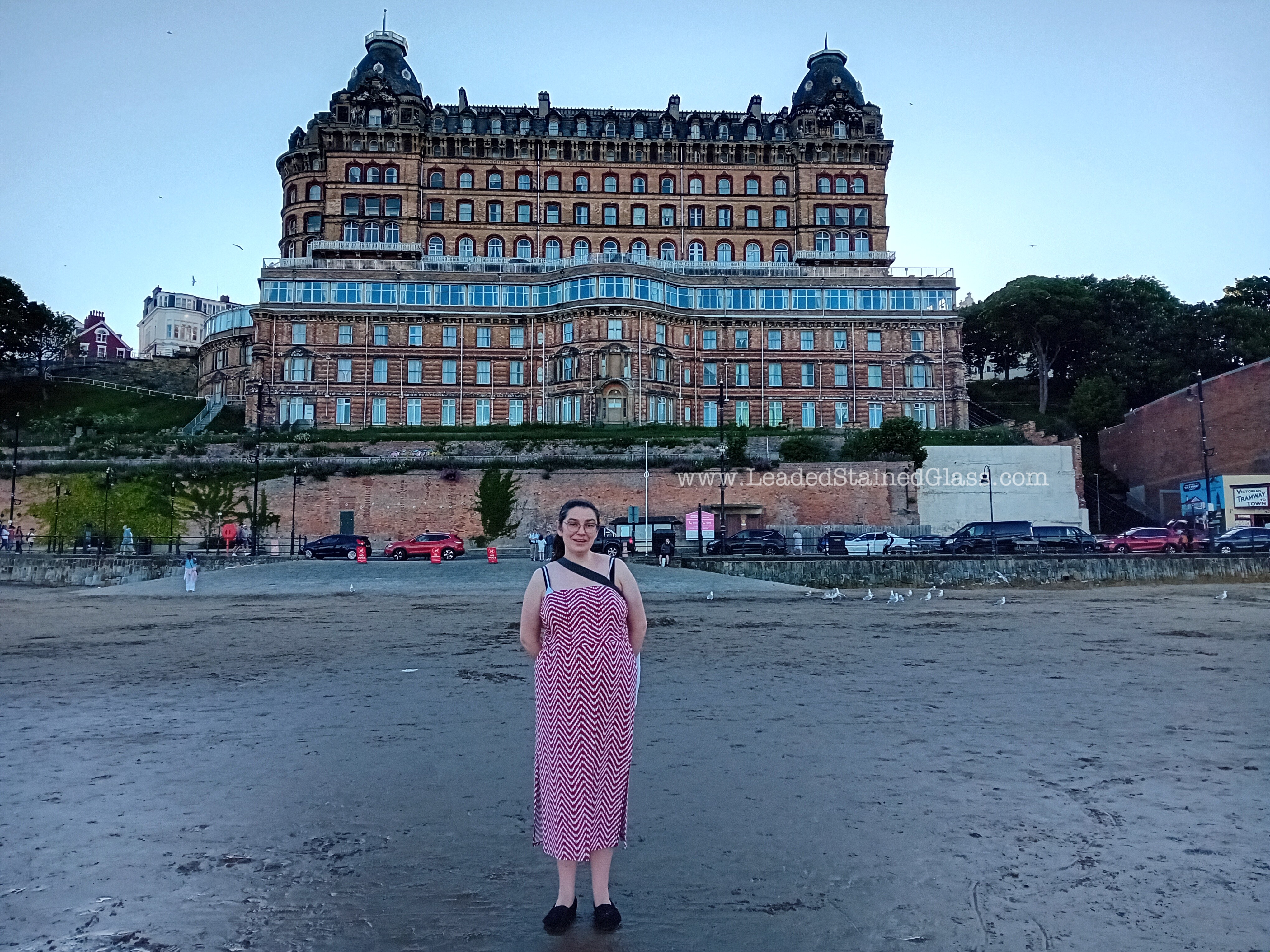 A woman in a long red dress and black shoes standing on Scarborough beach. In the background is the back of the Grand Hotel, cars and seagulls.